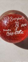 Top-down view of clear spherical paperweight with red core and white vintage Coca-Cola print including text and woman holding glass.