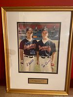 Full view of framed autographed photo of Tom Glavine and Greg Maddux in Atlanta Braves uniforms on a baseball field, with gold frame and white matting.