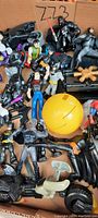Close-up photo showing various superhero and action figures including Batman figures, large yellow ball, and assorted toy accessories inside a cardboard box.
