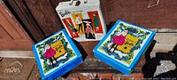 Photo of all three vintage vinyl Barbie doll carry cases displayed outdoors on a wooden surface, showing one white case and two blue cases with pink dress Barbie illustrations.