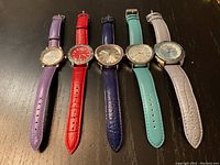 Front view of five ladies watches with colored leather straps laid out side by side, showing full bands and watch faces