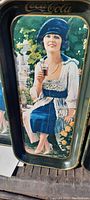 Close view of one Coca-Cola metal advertising tray showing woman holding a glass of soda with blue dress and floral background