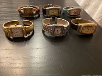 Set of six ladies cuff style watches arranged in two rows on a black surface, showing varied band materials and face shapes.