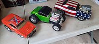 Four die cast vehicles including an orange Camaro with Confederate flag and '01' markings, green hot rod with exposed engine, and a patriotic American flag themed car on a white surface