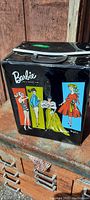 Close-up of one side of the black vinyl Barbie doll carry case showing colorful illustrations of Barbie in various outfits and 'Barbie' branding.