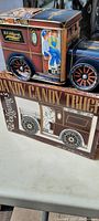 Photo showing side and top views of the colorful Wolfgang Candy Co. Dandy Candy Truck tin container on top with original box below. The tin features painted illustrations of children sitting on the truck and stylized wheels.