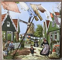 Front view of the Dutch hand decorated ceramic tile showing a lively street and village scene including traditional houses, laundry hanging on a line, villagers in costume, and a windmill.