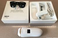 Photo showing the IPL device, sunglasses, razor, power cord, and original box arranged on a surface.