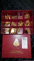 Full view of open red display box containing 12 compartments each holding gold-plated Christmas ornaments, with the box lid showing Danbury Mint logo and gold Christmas tree design.