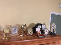 Wide angle showing the full assortment of eight clocks arranged on a wooden shelf against a tan wall.