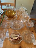 Overview of assorted cut glass bowls, dishes, and serving trays displayed on wood table, including amber bowl and two-tier tray with handle.