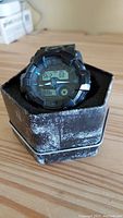 Front view of black G-Shock watch in worn black tin box on wooden surface.
