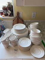 Photo showing stacked white ceramic dishes including plates, bowls, and mugs alongside several wood cutting boards with handles on a kitchen counter.