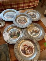 Set of 6 pewter plates on a wooden surface showing various colored battle scenes from the American Revolution with 'THE GREAT AMERICAN REVOLUTION 1776' engraved on rims