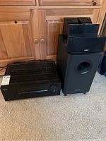 General view of AV receiver unit and subwoofer placed on carpeted floor near wooden cabinet.
