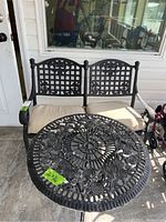 Front view of black metal patio loveseat with beige cushions and round metal table with ornate floral design and size label 26x26.
