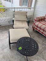 Photo showing the rocker lounge chair with cushions, ottoman with matching cushion, and metal patio table arranged on porch floor.