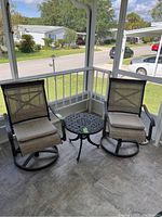 View of two metal swivel rocker patio chairs with beige cushions and round metal table between them on enclosed porch.