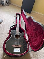 Rogue Model AB 304 A acoustic bass guitar resting in a fitted hard case with plush red interior lining. Guitar has black glossy finish and natural wood bridge.