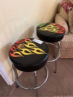 Two vintage bar stools with black flame patterned tops and chrome legs against a beige wall and carpeted floor.