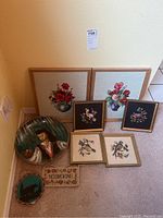 Photo showing full view of assorted embroidered artworks and ceramic wall pieces arranged on carpeted floor against wall.