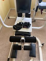 Front view of the gym system bench area with padded seat, leg support pads, and a pair of black workout gloves laying on the seat.