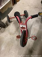 Full view of the red antique Lakeshore steel tricycle with rubber wheels on a concrete floor.