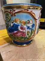 Close up of hand painted raised relief scene on vase showing classical figures, floral motifs and gold trim.