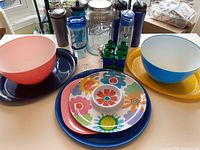 Photo showing handmade plastic serving trays with colorful flower designs stacked on dark blue trays, large pink and blue plastic bowls, assorted water bottles, and a clear glass lemonade dispenser with a metal lid at the back.