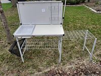 Camping kitchen set up outdoors on grass showing sink, folding shelves and storage racks.