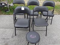 Front view of 4 black folding chairs and 1 round folding stool arranged outdoors on pavement.