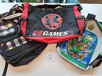 Photo showing the two backpacks side by side on a table. The EB Games tote is black with red trim and a large EB Games logo with a video game controller. The T-Rex backpack is green with a colorful skateboard-riding dinosaur graphic and mesh side pockets.