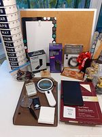 Photo showing cork bulletin board, perpetual calendar, clipboard with notepad, magnifying glasses, Panasonic battery charger, new sealed laptop lock and WiFi adapter, plus various smaller office items on white table.