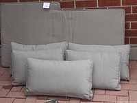 Six grey outdoor cushions against a brick wall, three larger seat cushions with notch detail and smaller back cushions arranged in front.