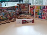 Five assorted 300 piece puzzles shown in their boxes on a windowsill with outdoor view, including Campside, autumn forest, donuts, popsicles, and boats themes.