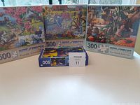 Four unopened jigsaw puzzle boxes showing vibrant animal and nature scenes on the covers, including a dog, deer, birds, raccoon, and garden settings. Quantity and piece count visible on each box.