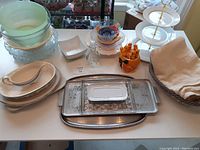 Front view showing a variety of serving dishes including glass bowls stacked, ceramic dishes including gravy boat, metal trays, small salt and pepper shakers, orange grapefruit set, and folded cloth napkins or towels.