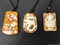 Photo showing all three ambergris pendants laid side by side on black textured surface, highlighting their yellow amber and white carvings.