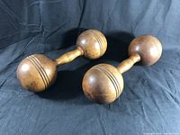 Pair of antique wooden dumbbells placed on black fabric background; showing carved grooves and smooth turned handles on both dumbbells.