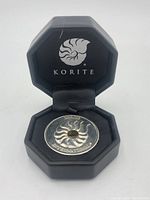 Silver-tone Korite ammonite coin with colorful ammolite gemstone center inside black octagonal display case with Korite logo