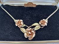 Close-up of the gold rolled floral necklace with three rose flowers and leaves on a black velvet cushion.