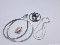 Two silver chains (flat herringbone and thin snake chain) laid out alongside two pendants (sun face and camel with abalone shell) on white background.