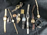 Top-down view of 11 vintage wristwatches of various styles and bands, displayed on black fabric.