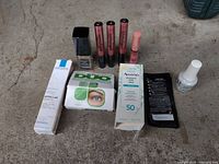 Overview of all 10 cosmetic items arranged on concrete floor showing skincare, foundation, lipsticks, sunscreen, lash adhesive, nail polish, and sample sachets.