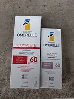 Photo of Garnier Ombrelle Complete Sensitive Advanced Lightweight Lotion SPF 60, 200 ml and Garnier Ombrelle Face Ultra-Light Hydrating Cream SPF 60, 75 ml, side by side on a grey concrete floor.