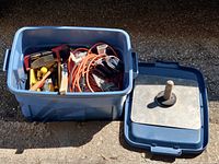Blue plastic storage bin containing assorted hand and power tools including a corded drill, extension cord, hammer, handsaw, level, clamps, and other small hand tools, as well as the tote lid placed to the side.