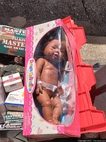 Photo showing the Dream Collection baby newborn doll, brown skin tone, in original pink box with plastic window, doll wearing white diaper, lying down inside box.