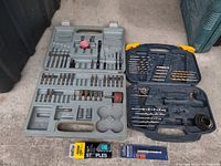 Two open plastic cases showing assorted drill bits and hole saws arranged inside, used and incomplete. New packs of staples visible below the cases.