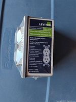 Box packaging showing Leviton brand, weather resistant kit, 15A 125V standard receptacle with U-ground duplex and contents listed in English and French.