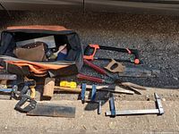 Photo showing Ridgid large 26-inch orange and black tool bag filled with assorted hand tools including saws, mallet, clamps, and shears laid out on ground.